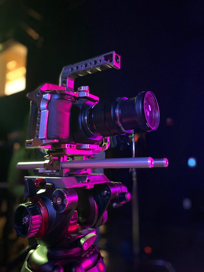 Close-up of a high-tech video camera mounted on a sturdy tripod, illuminated by vibrant pink lighting.